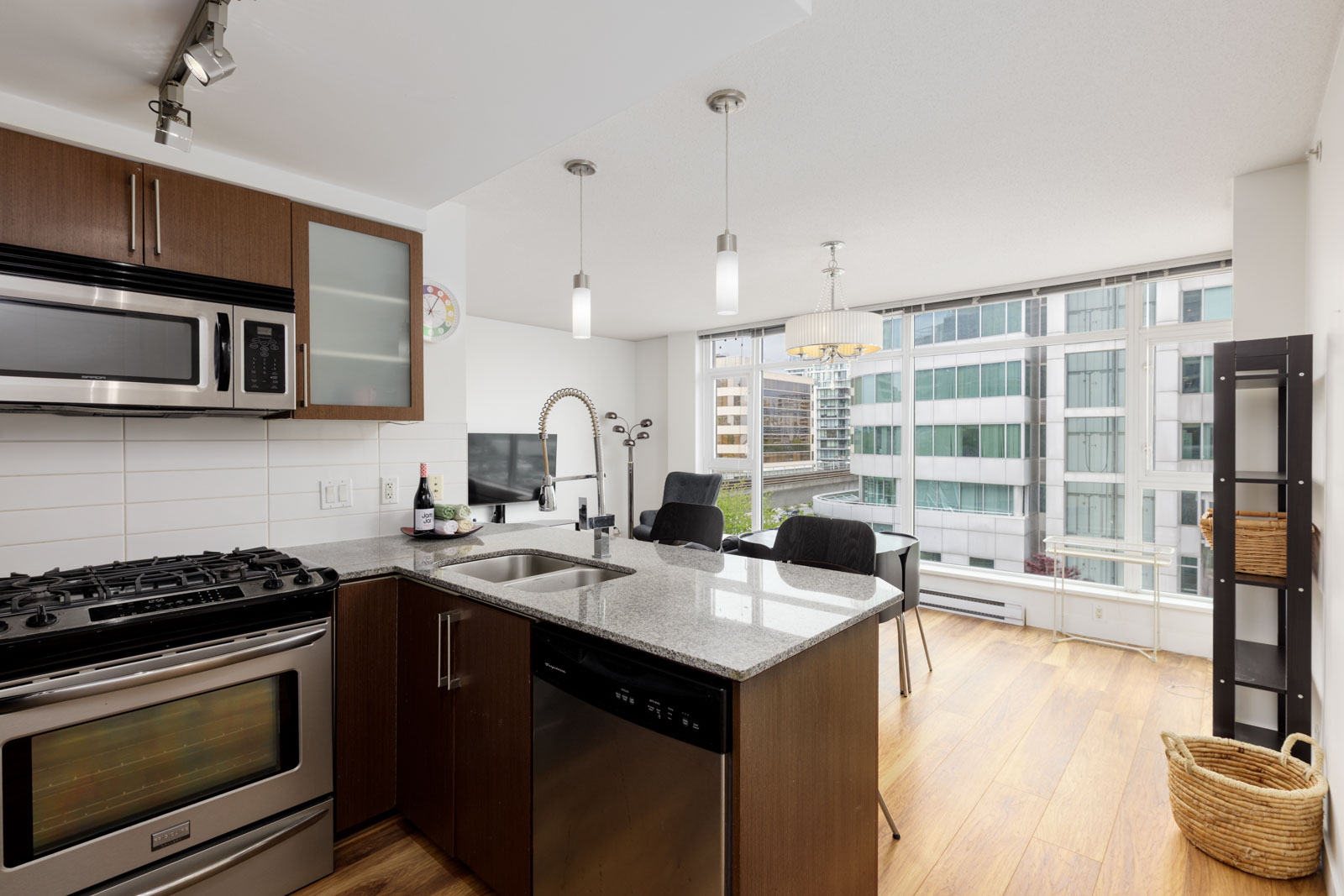 Modern apartment kitchen with stainless steel appliances, wood cabinets, an island sink, and adjacent dining area next to large floor-to-ceiling windows overlooking city buildings.