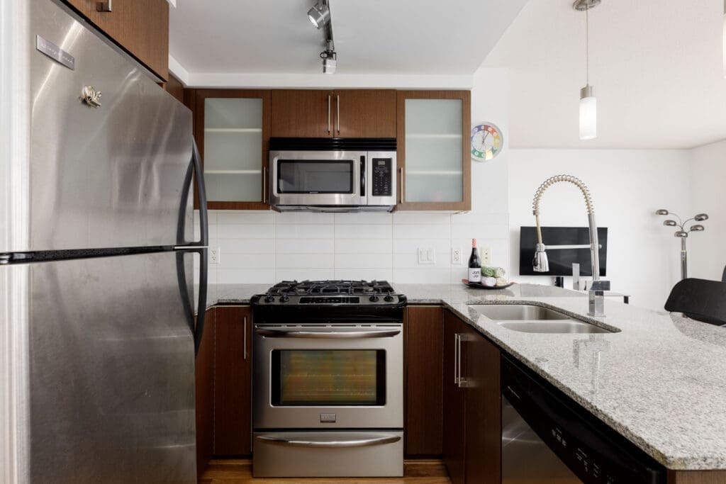 Modern kitchen with stainless steel appliances, a granite countertop, and wood cabinets; wine bottles and glasses are placed near the sink.