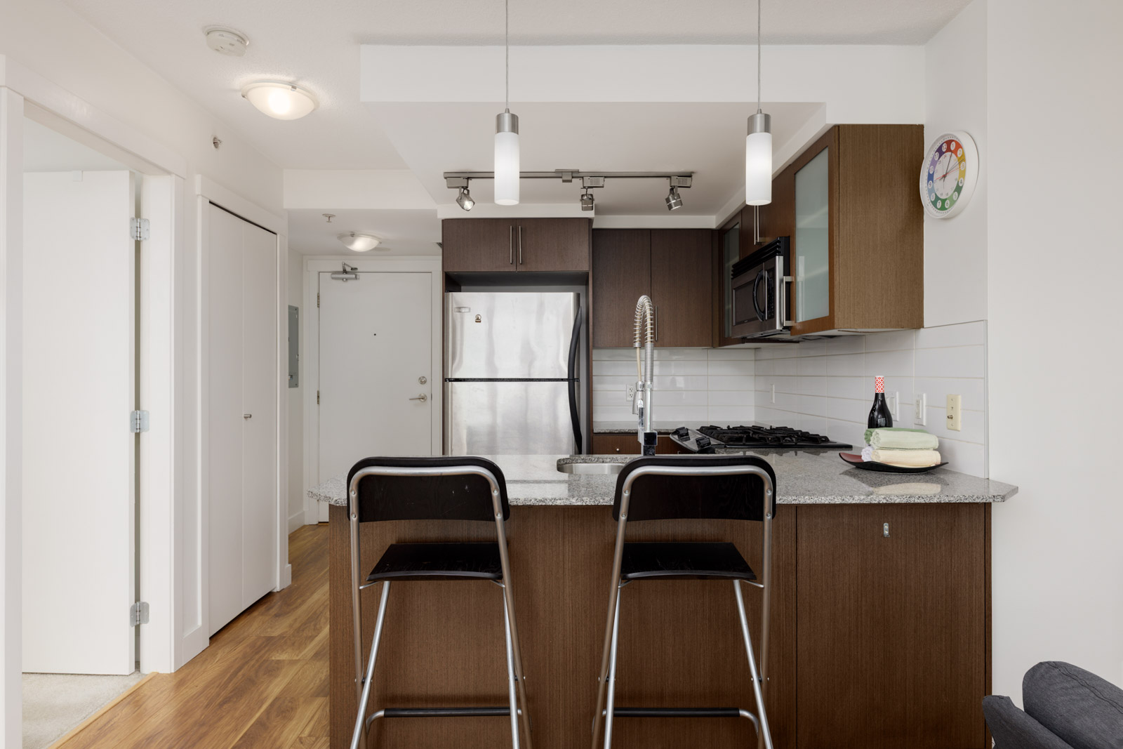 Modern kitchen with stainless steel appliances, dark wood cabinets, a granite island with two bar stools, pendant lighting, and a bottle of wine on the counter.