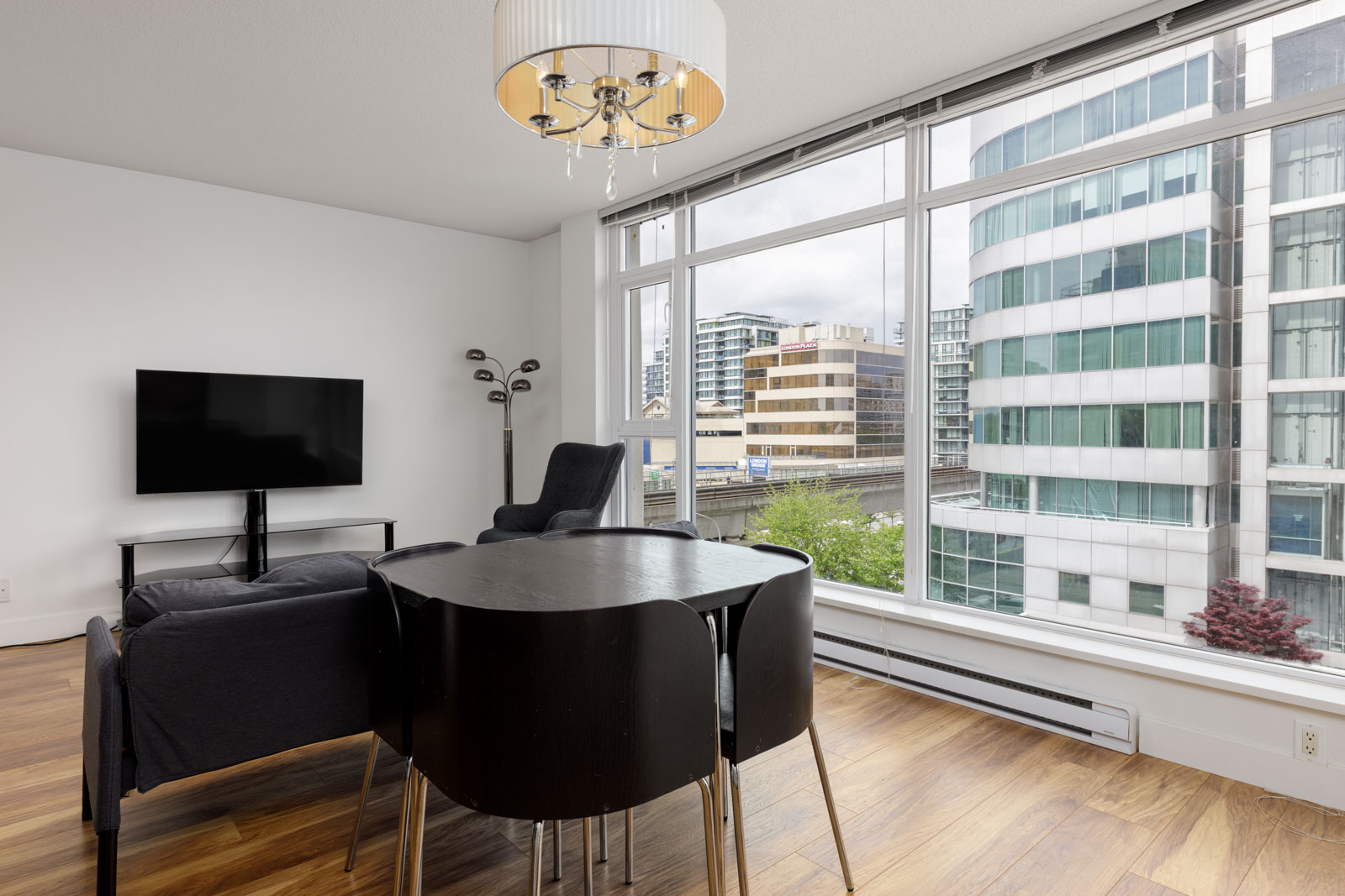 Modern living room with a black dining table, dark sofa, TV, large window, and city building views. Wood flooring and minimal decor.