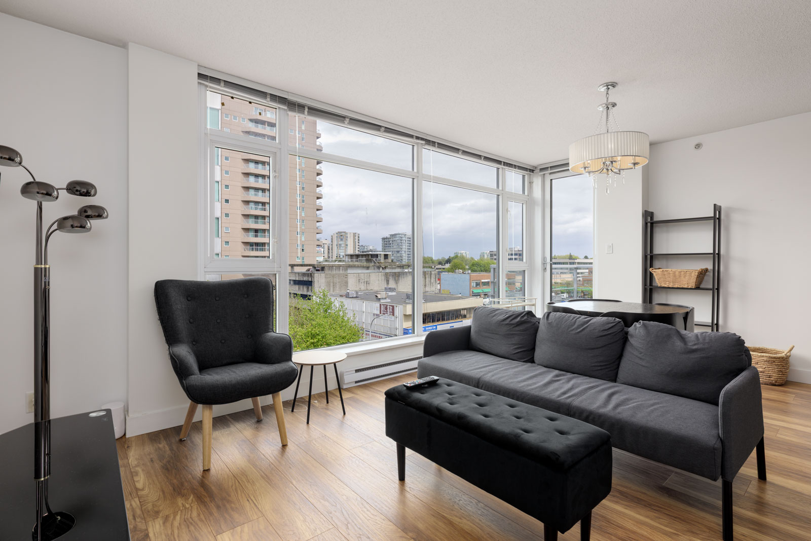 Modern living room with a black sofa, armchair, ottoman, floor lamp, dining table, and large windows overlooking city buildings and greenery.
