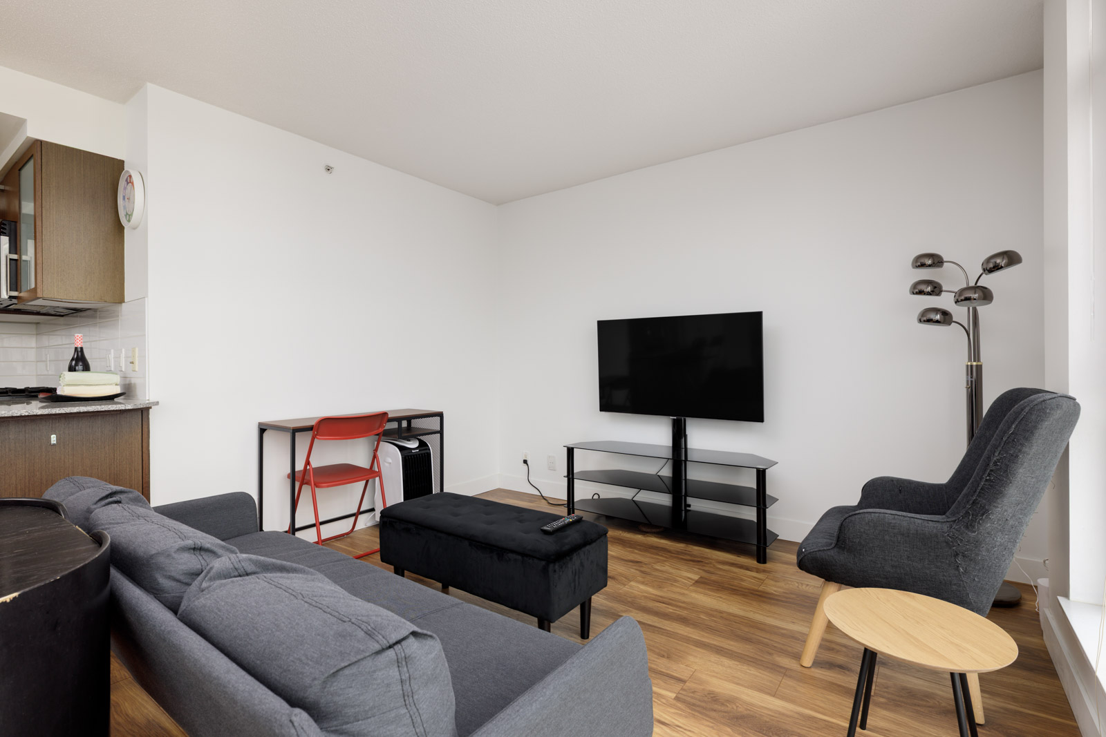 Modern living room with a gray sofa, armchair, wall-mounted TV, black TV stand, red desk with a chair, floor lamp, and wood flooring. Kitchen area visible on the left.