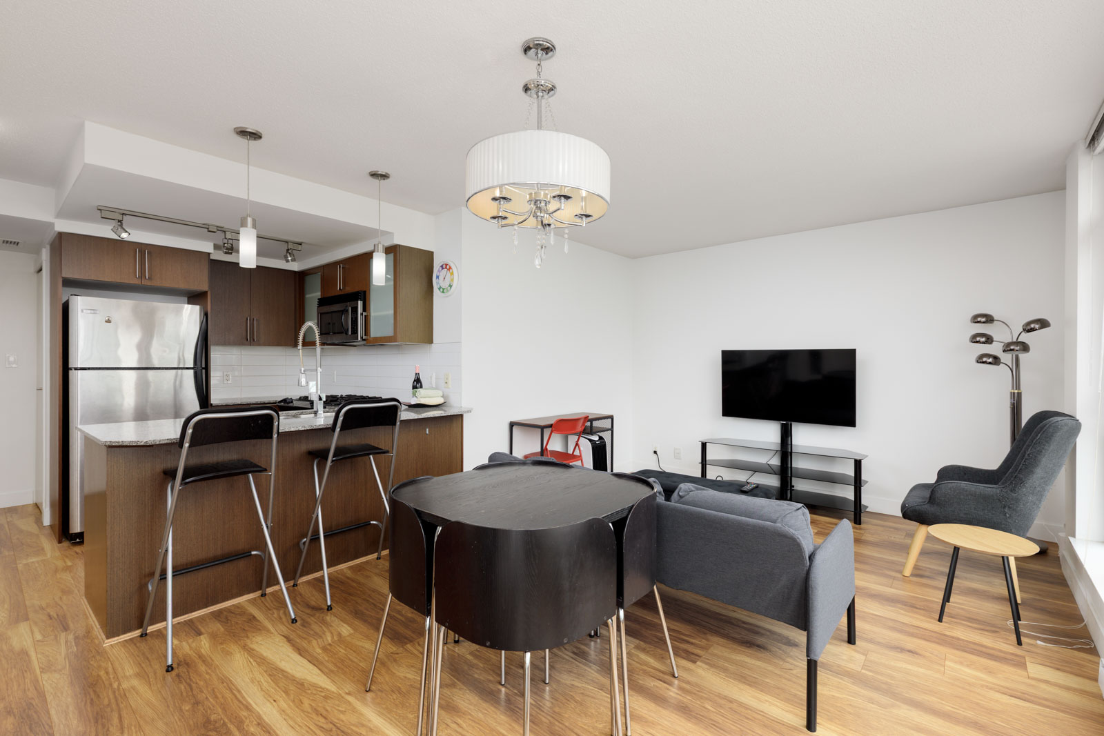 Modern open-plan living room and kitchen with wood floors, dark furniture, stainless steel appliances, a wall-mounted TV, and large windows.