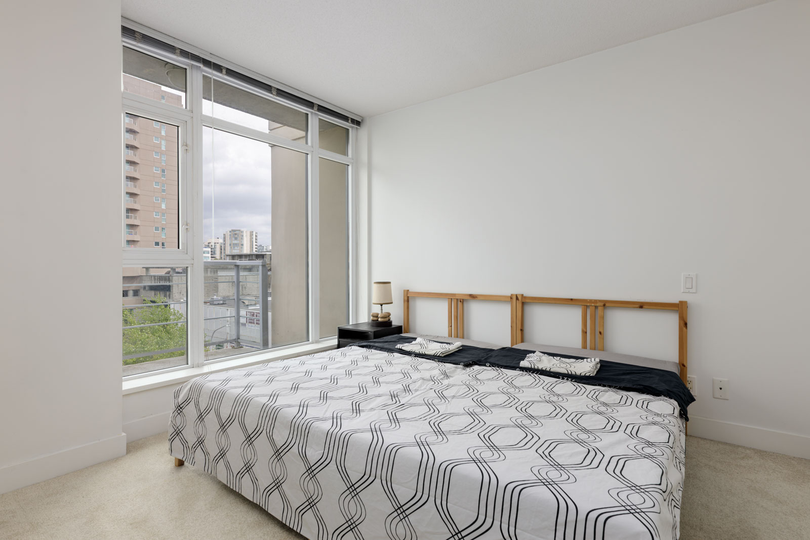 Minimalist bedroom with a large window, white walls, a double bed with a patterned bedspread, and two small bedside tables. Urban buildings visible outside.
