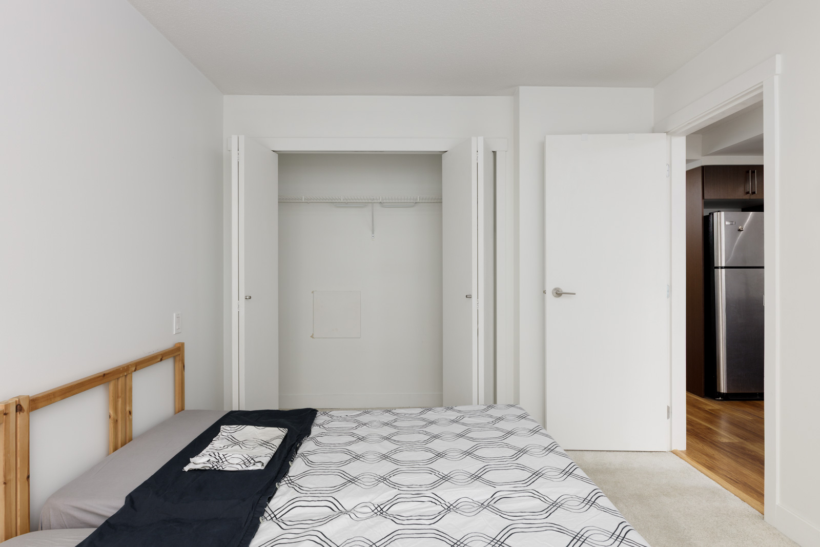 A minimalist bedroom with an unmade bed, open closet with no items inside, white walls, and a view of a kitchen with a stainless steel refrigerator.
