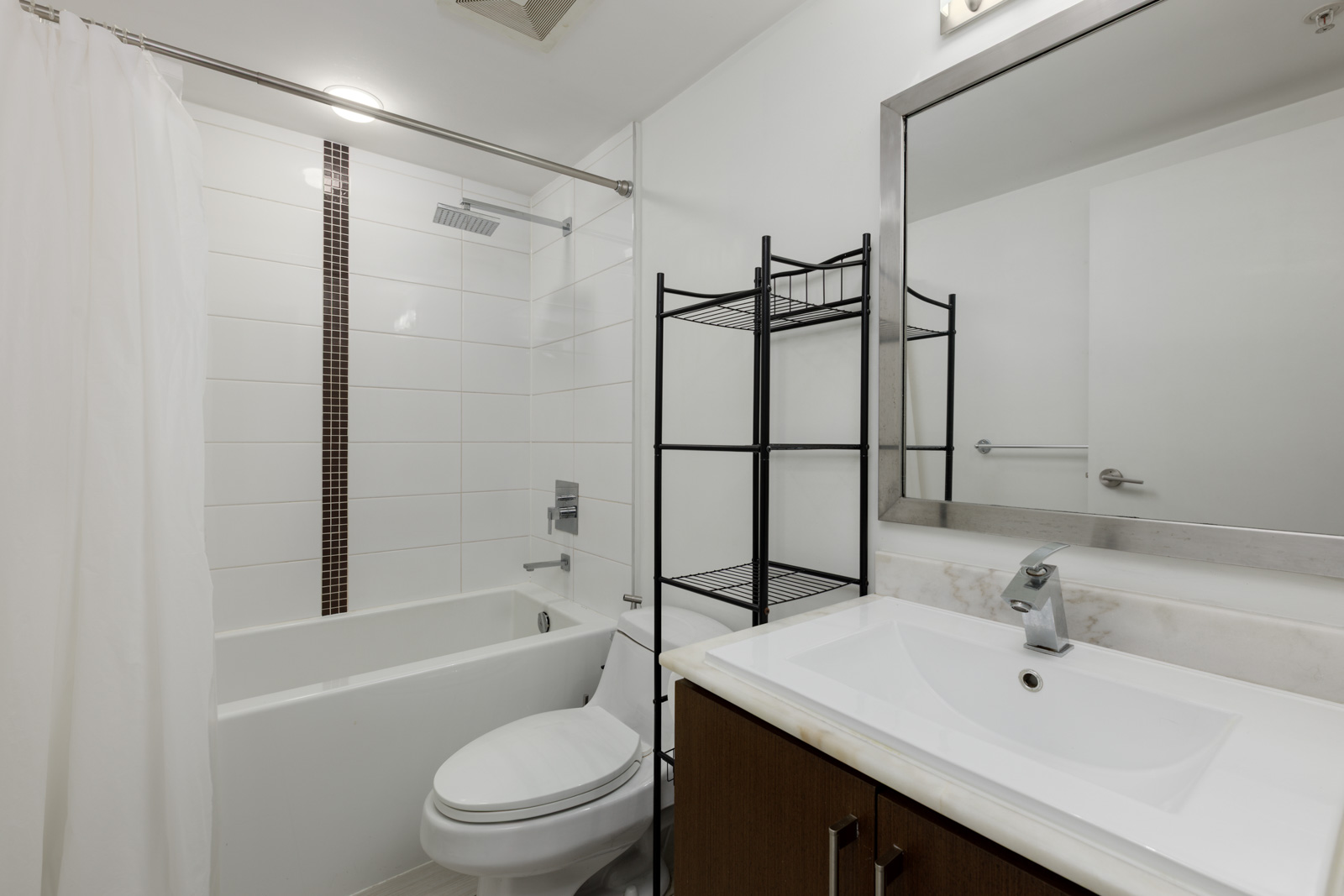 Modern bathroom with white tile walls, a bathtub with shower, toilet, black metal shelving unit, and a sink with a rectangular mirror above it.