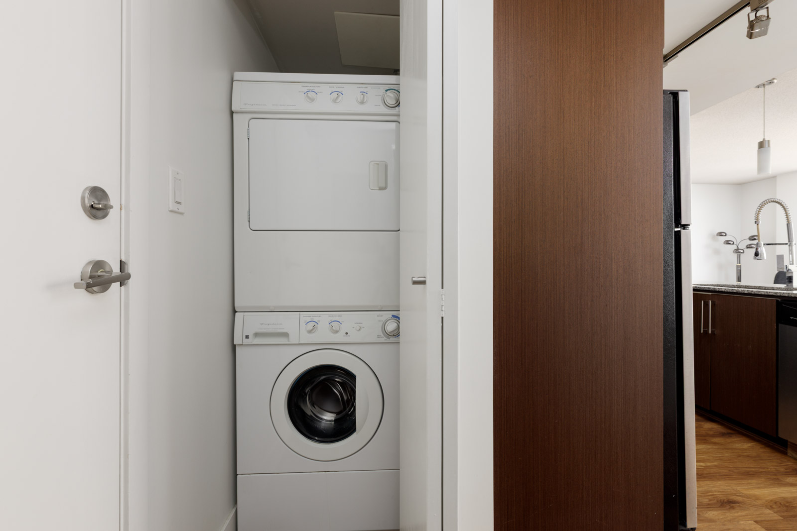 A stacked washer and dryer unit is placed in a small closet next to a kitchen area in a modern apartment.