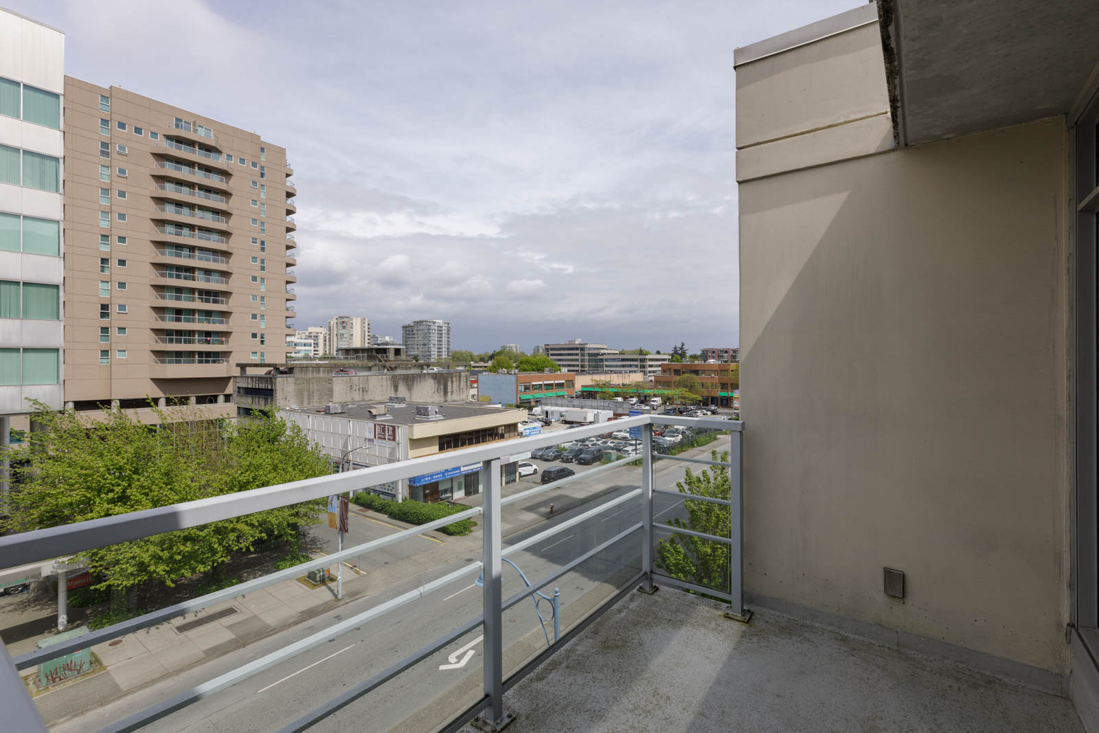 View from a balcony overlooking a city street with mid-rise buildings, trees, parked cars, and a partly cloudy sky.