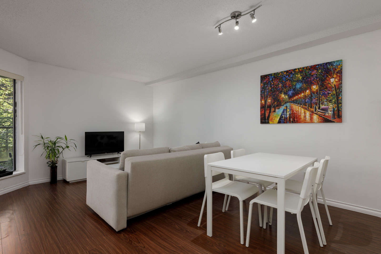 A modern living and dining area with a beige sofa, white dining table with four chairs, wall-mounted TV, floor lamp, large window, and a colorful painting on the wall.