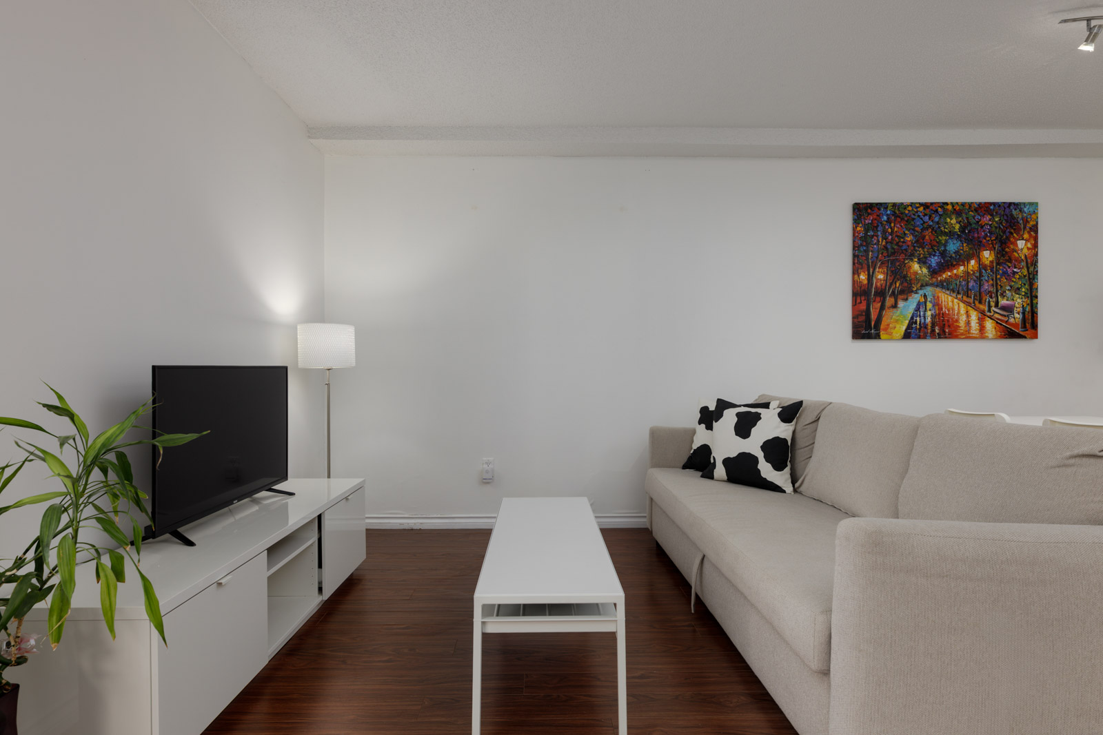 A modern living room with a beige sectional sofa, a white coffee table, a TV on a white stand, a floor lamp, a potted plant, and a colorful painting on the wall.