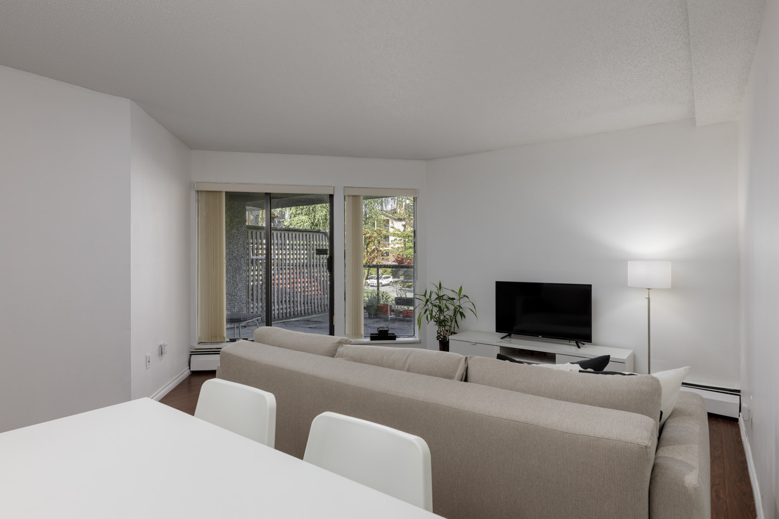 A living room with a light gray sofa, a TV on a stand, a floor lamp, a potted plant, and sliding glass doors opening to a patio area with outdoor plants.