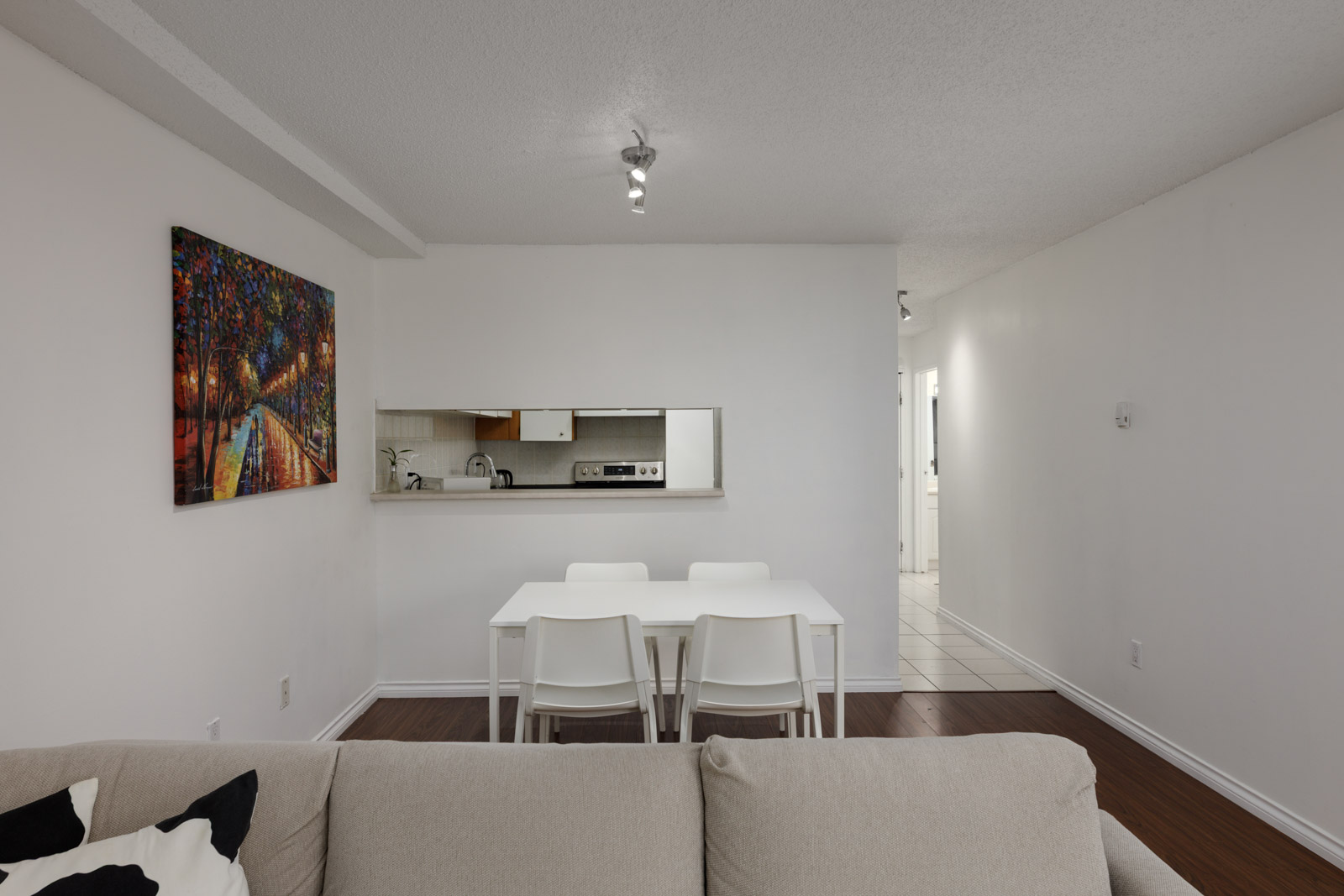 A modern dining area with a white table and four chairs, adjacent to a small kitchen with a pass-through window, white walls, and a colorful painting on the left wall.
