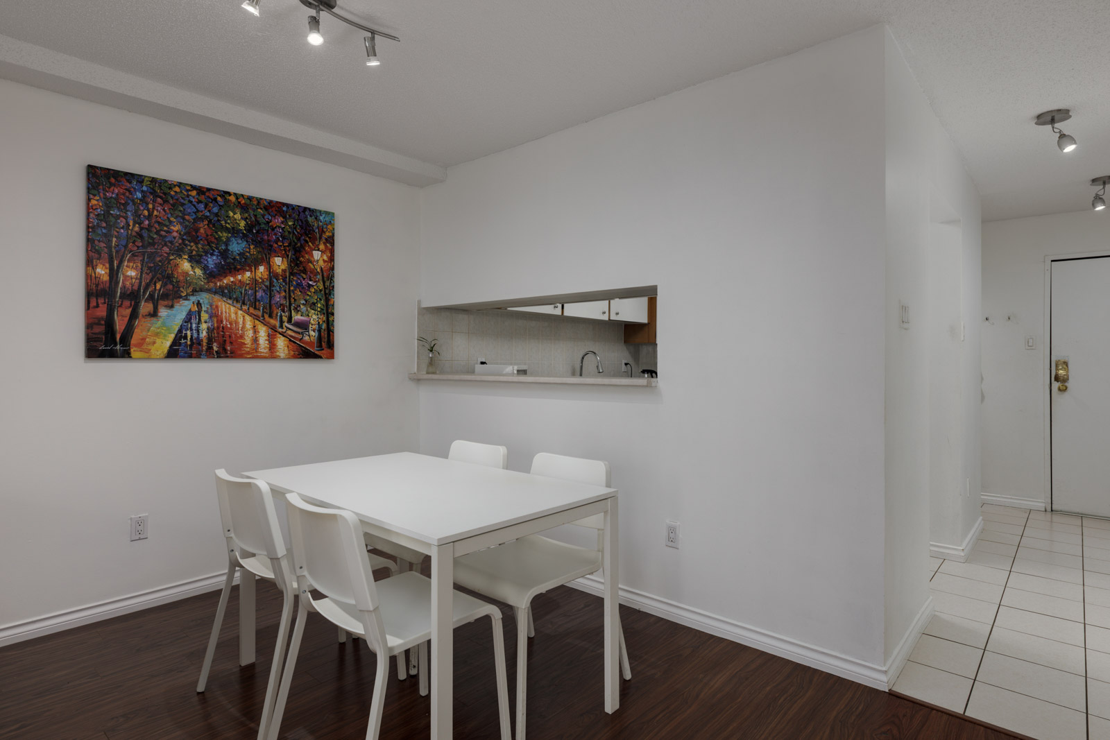 A small dining area with a white table and four chairs, a colorful painting on the wall, and a kitchen visible through a serving hatch.