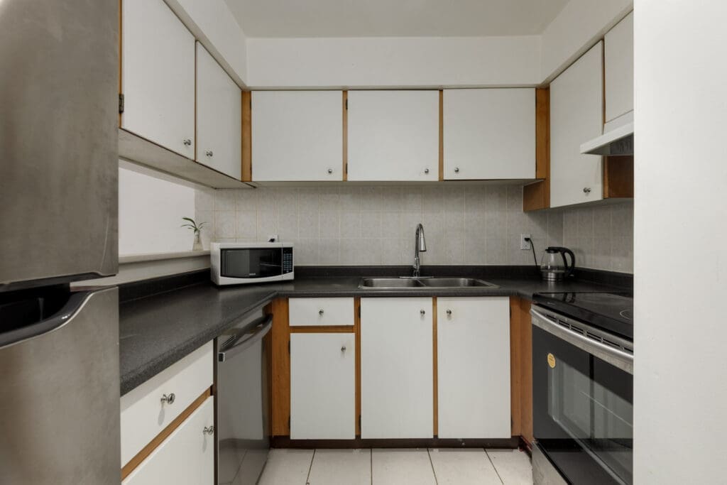 A small kitchen with white cabinets, black countertops, a sink, a microwave, a stove, a dishwasher, and a stainless steel refrigerator.