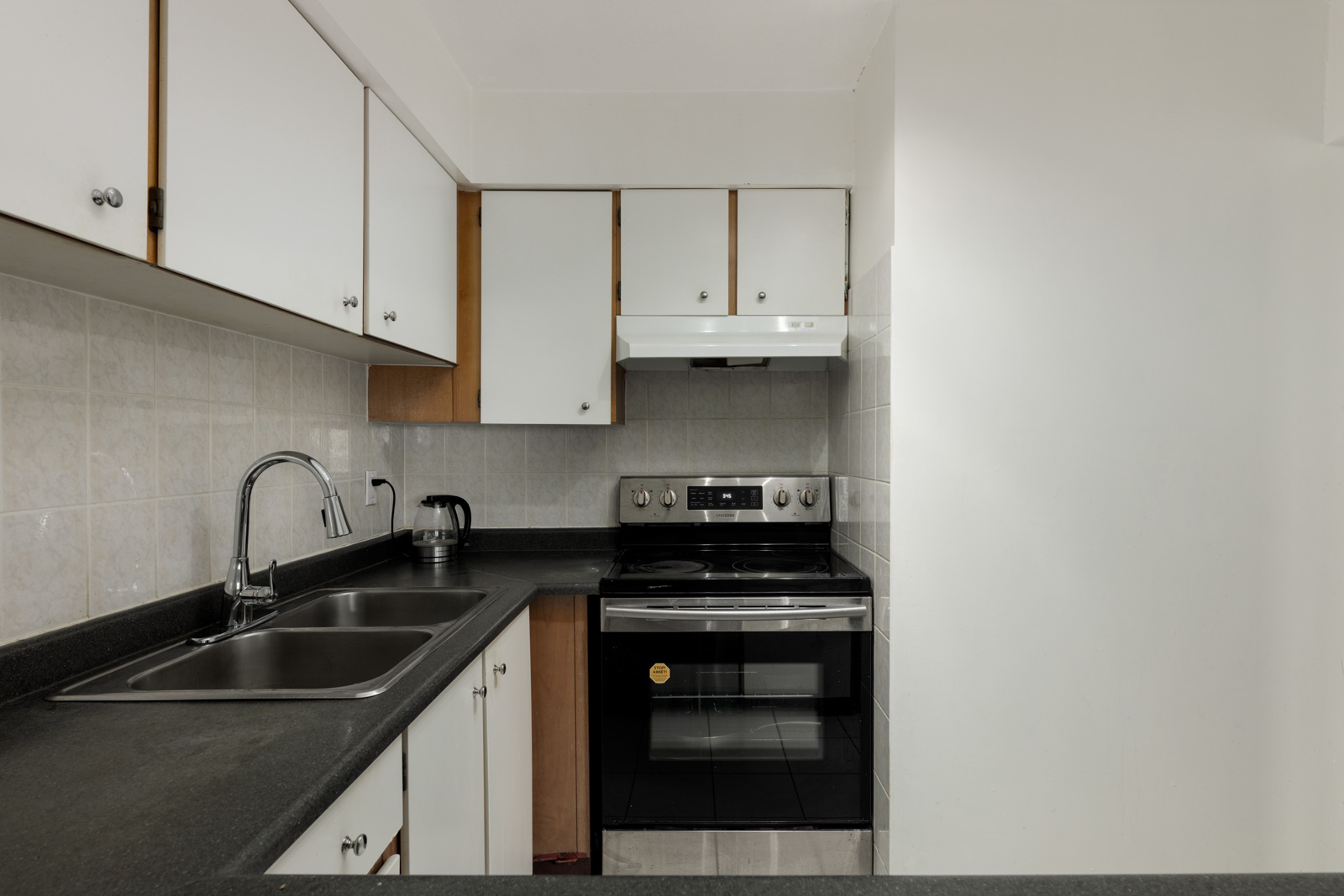 A compact kitchen with white cabinets, black countertops, a double sink, electric stove with oven, range hood, kettle, and tiled backsplash.