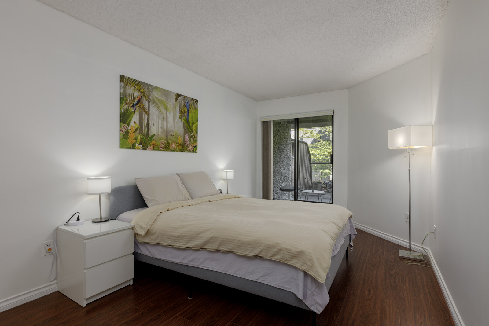 A bedroom with a double bed, two bedside tables with lamps, a floor lamp, wall art above the bed, and glass doors leading to an outdoor area.