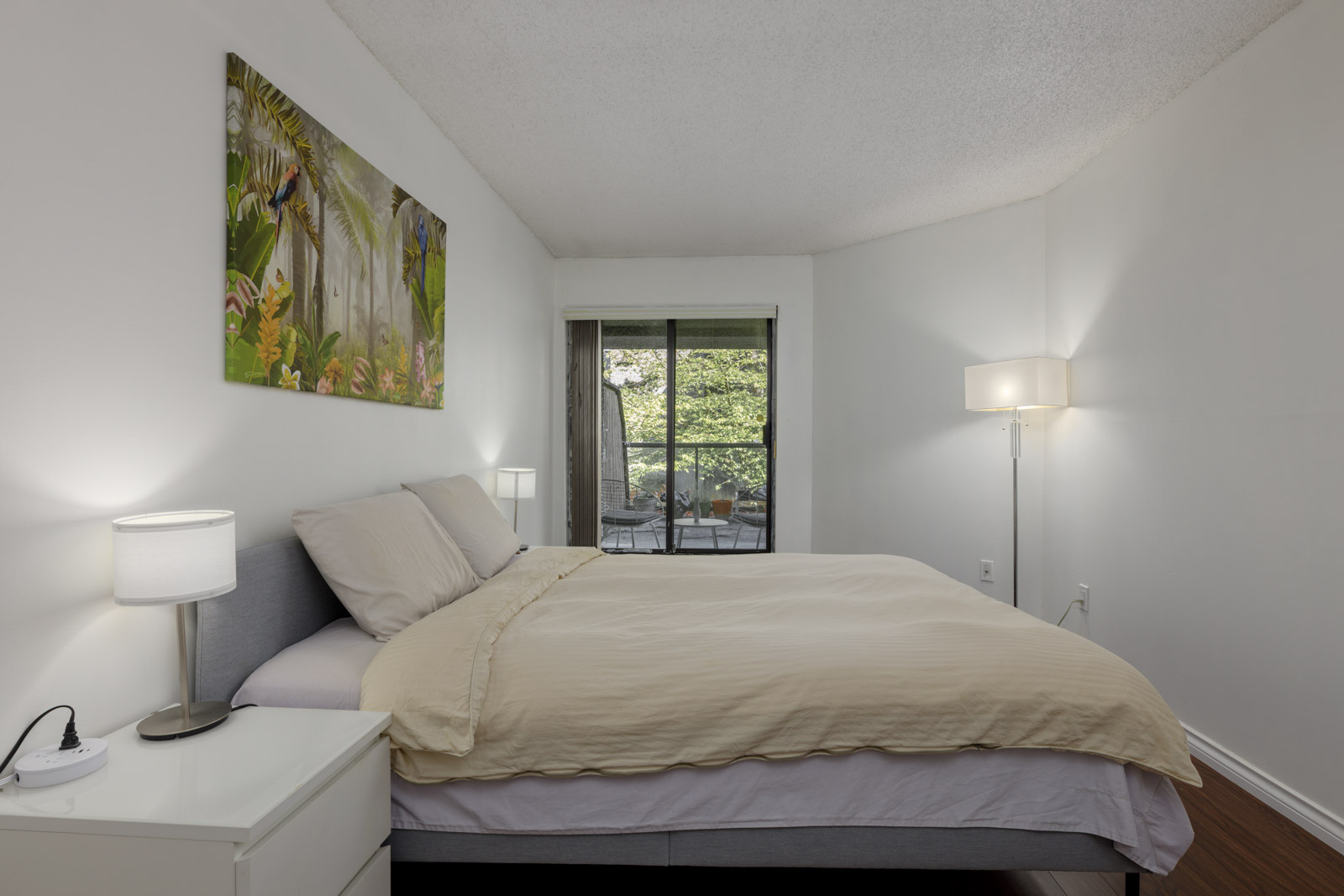A neatly made bed with beige bedding in a minimalist bedroom, two bedside lamps, wall art above the bed, and a sliding glass door leading to a balcony with greenery outside.