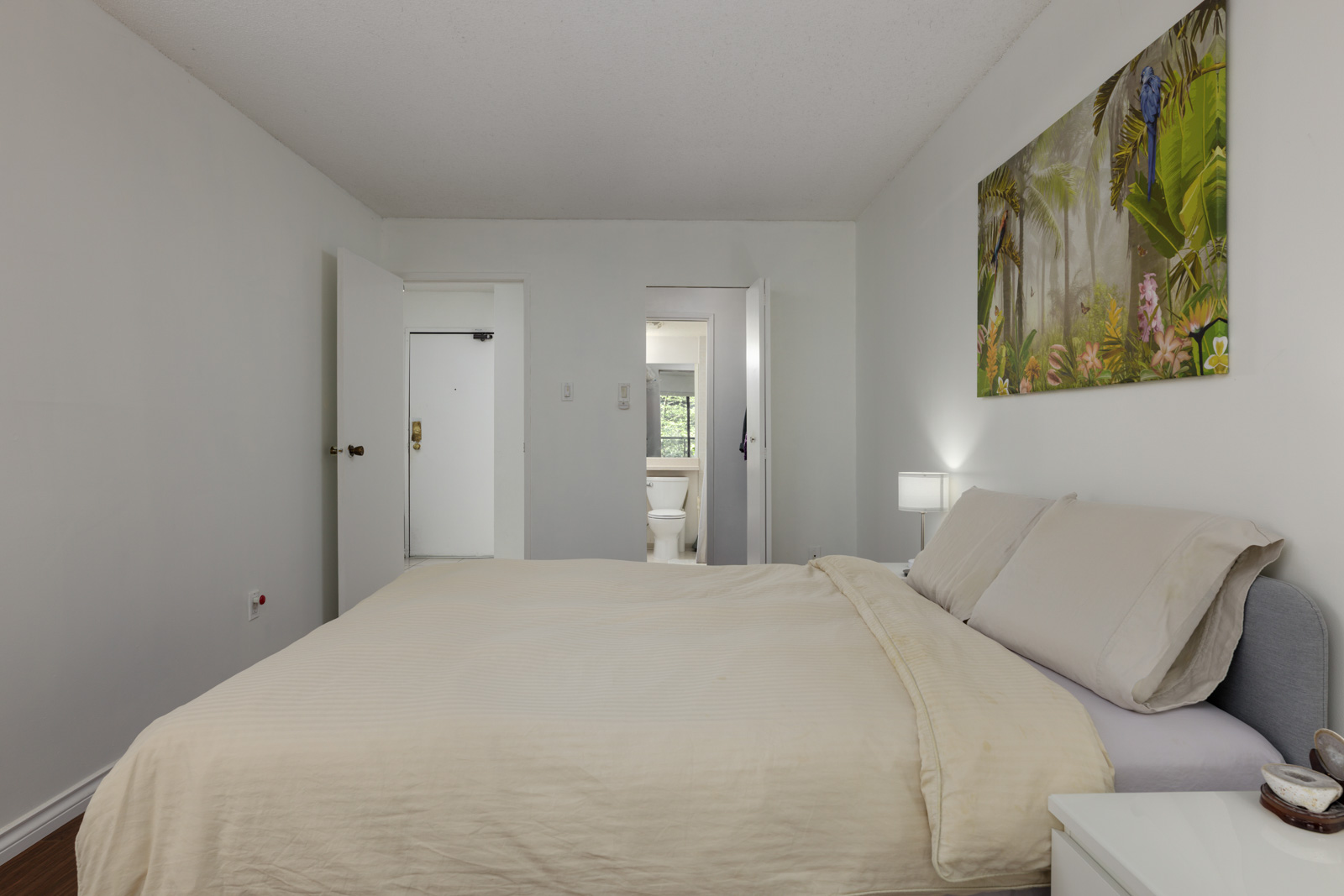 A bedroom with a large bed, light-colored bedding, a nightstand with a lamp, wall art, and an open door leading to a bathroom with a visible toilet.