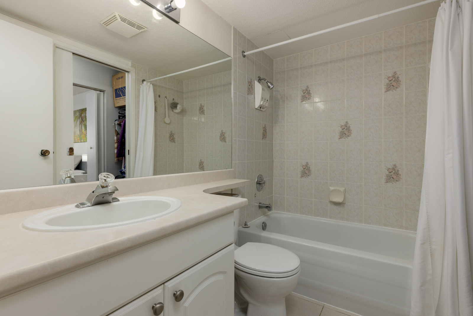 A small bathroom with a white sink, countertop, toilet, and bathtub with a shower curtain. Tile walls have decorative floral accents. A large mirror is above the sink.