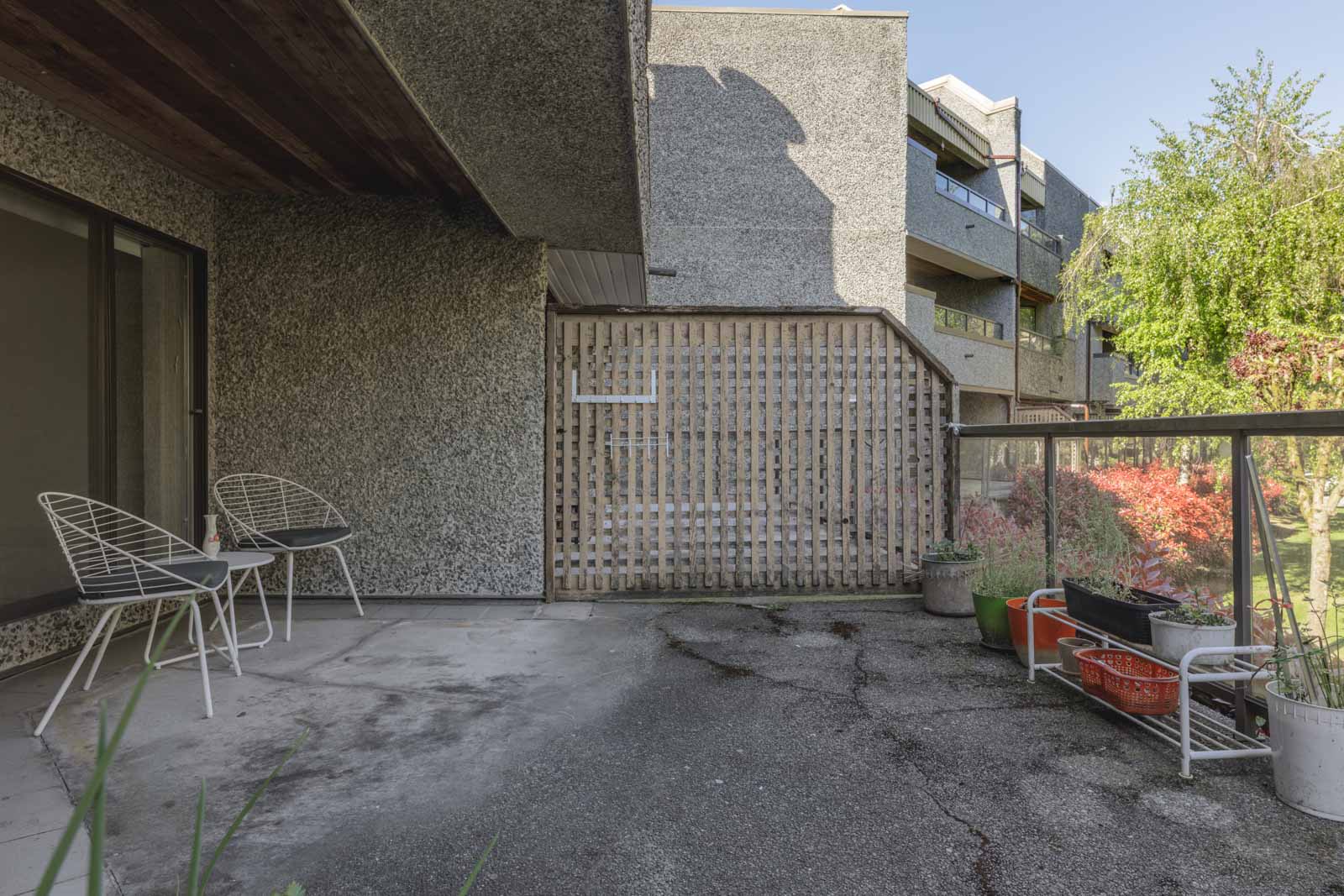 A spacious outdoor patio with two white chairs, a glass railing, potted plants, and a wooden privacy screen, adjacent to a concrete building.