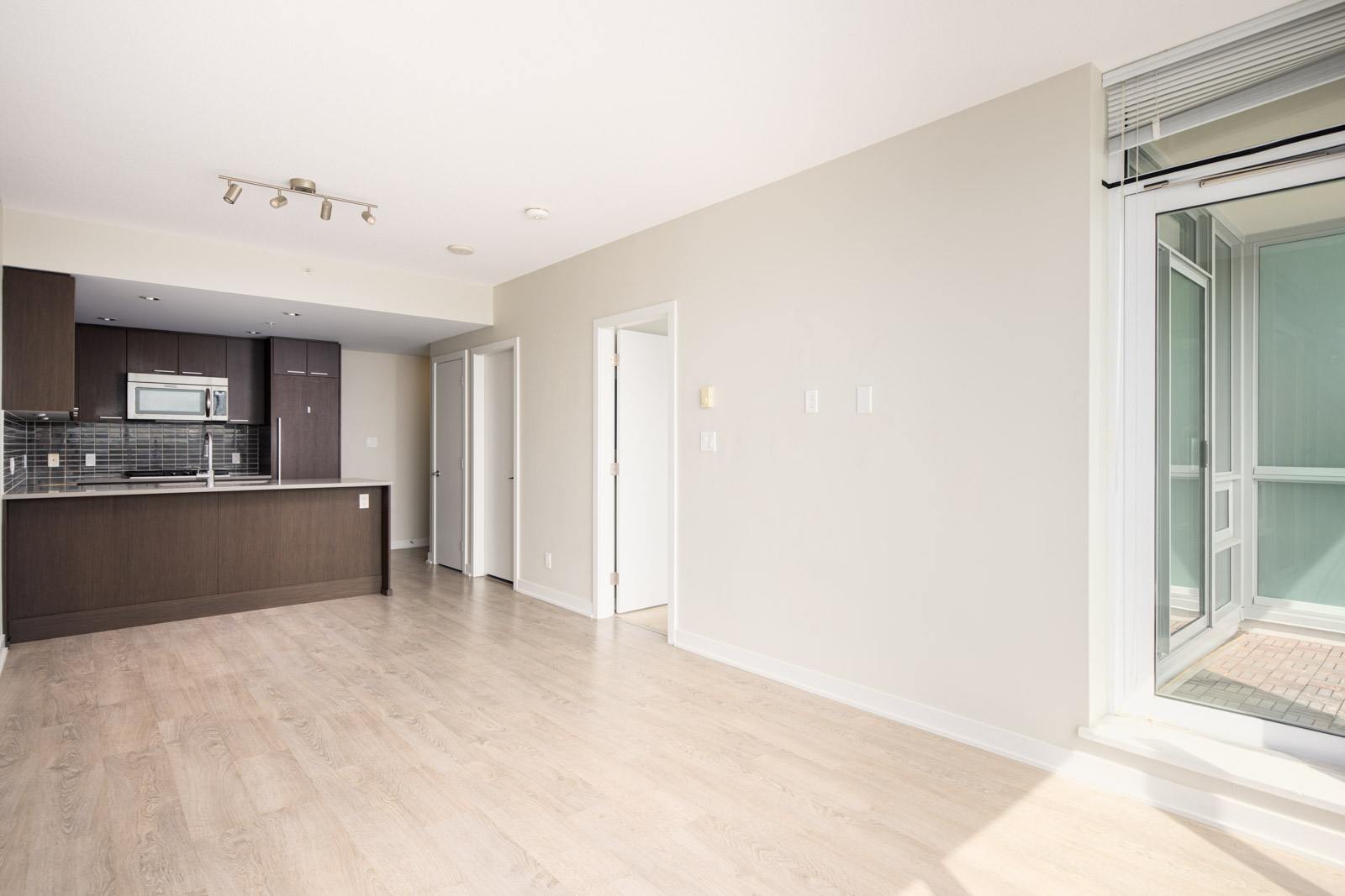 Bright, modern apartment interior with light wood floors, open kitchen with dark cabinets, and sliding glass door to a balcony.