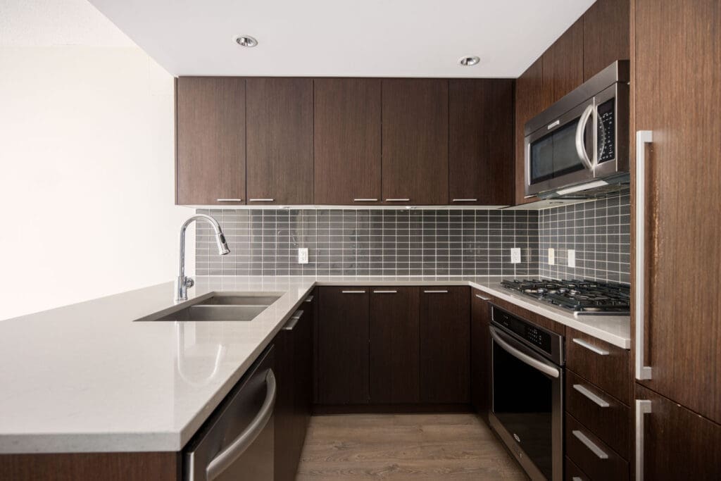 Modern kitchen with dark wood cabinets, stainless steel appliances, grey tile backsplash, white quartz countertops, and a built-in sink.