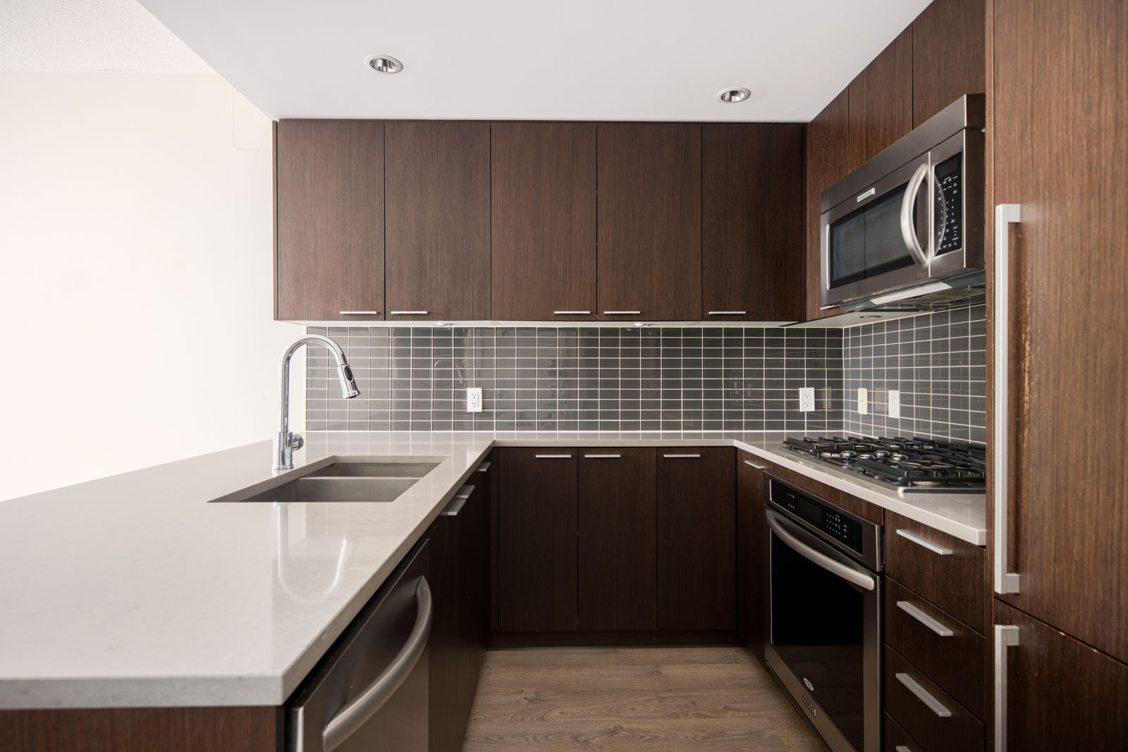 Modern kitchen with dark wood cabinets, stainless steel appliances, grey tile backsplash, white quartz countertops, and a built-in sink.
