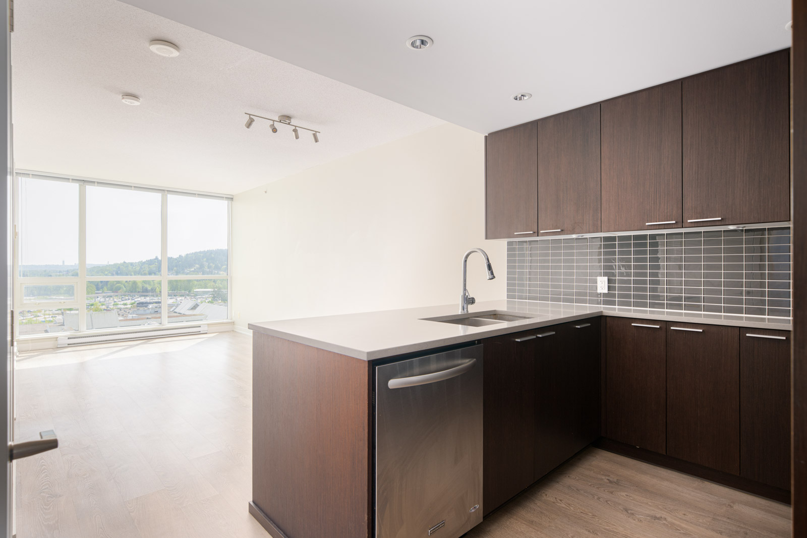 Modern kitchen with dark wood cabinets, gray tile backsplash, stainless steel appliances, and an open layout overlooking a bright living area with large windows.