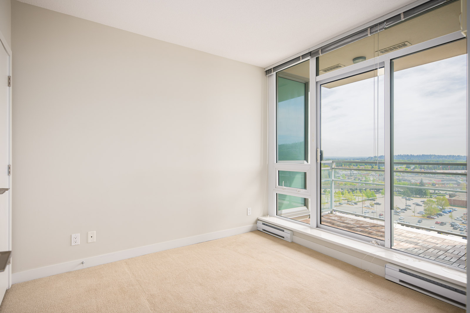 Empty room with beige carpet, light-colored walls, and large windows leading to a balcony with a view of a parking lot and greenery outside.