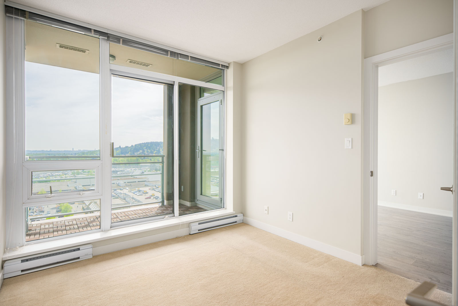 Bright, empty room with beige carpet, large floor-to-ceiling window, glass door to a balcony, and an adjacent room with hardwood flooring.