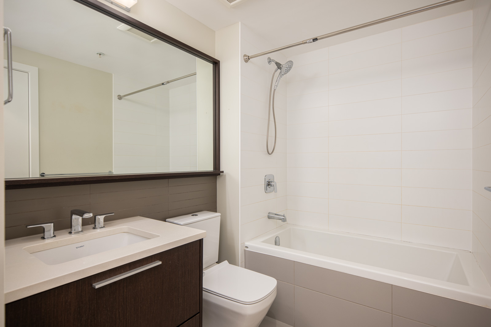 Modern bathroom with a rectangular sink, dark wood vanity, toilet, and a bathtub-shower combo with a handheld showerhead against white tiled walls.