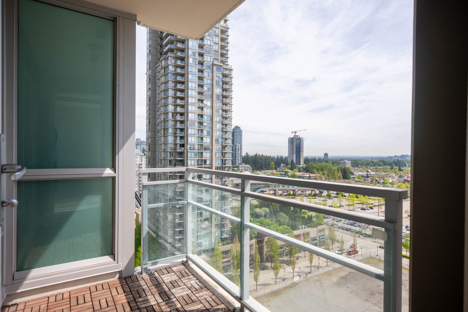 View from a balcony with glass railing, overlooking tall buildings, a construction crane, and a cityscape under a partly cloudy sky.