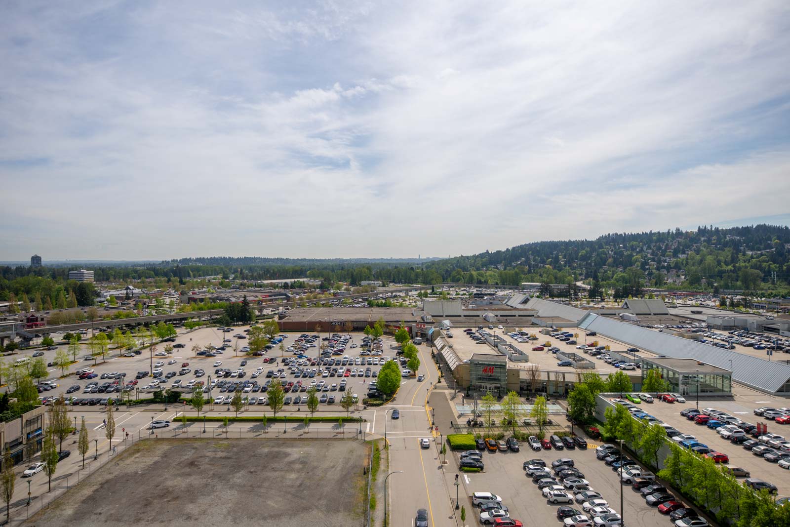 A large shopping center with multiple parking lots filled with cars, surrounded by trees and distant hills under a cloudy sky.