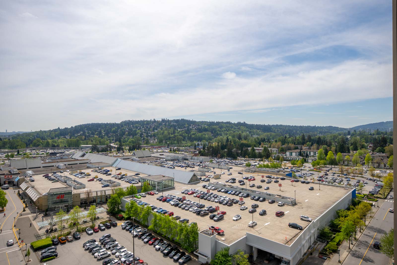 Aerial view of a large shopping center parking lot filled with cars, surrounded by green trees and hills under a partly cloudy sky.