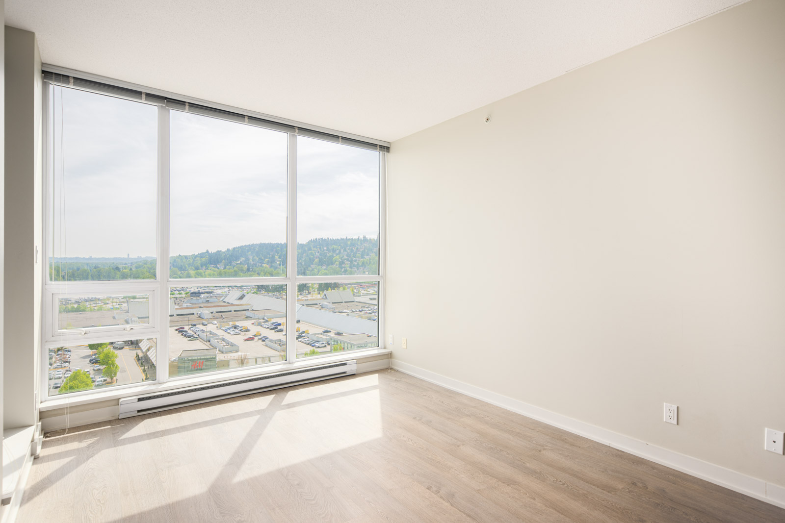 Bright empty room with large floor-to-ceiling windows, light wood flooring, neutral walls, and a view of an industrial area and greenery outside.