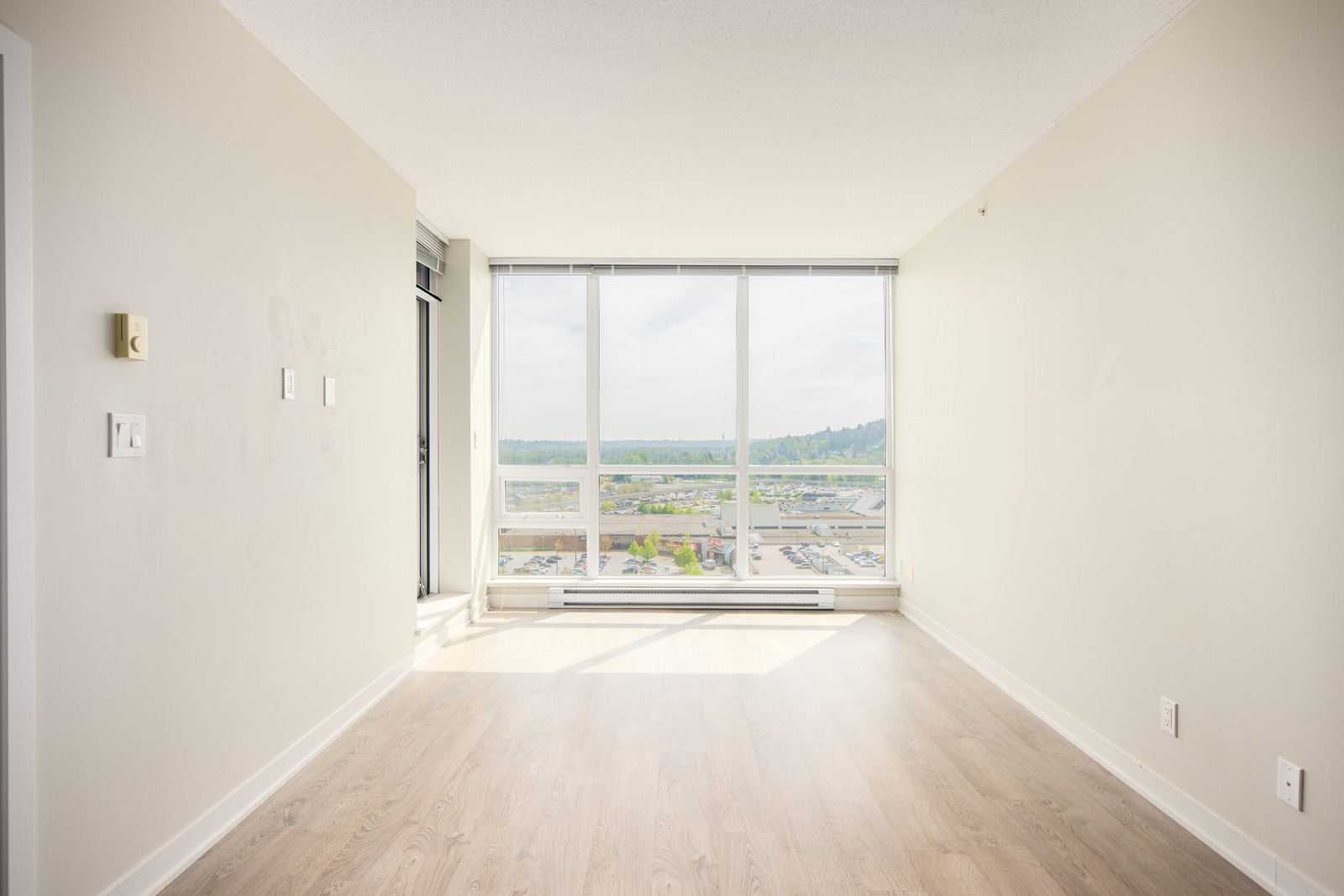 Empty room with light-colored walls, wood flooring, and large floor-to-ceiling windows overlooking a cityscape and distant hills.