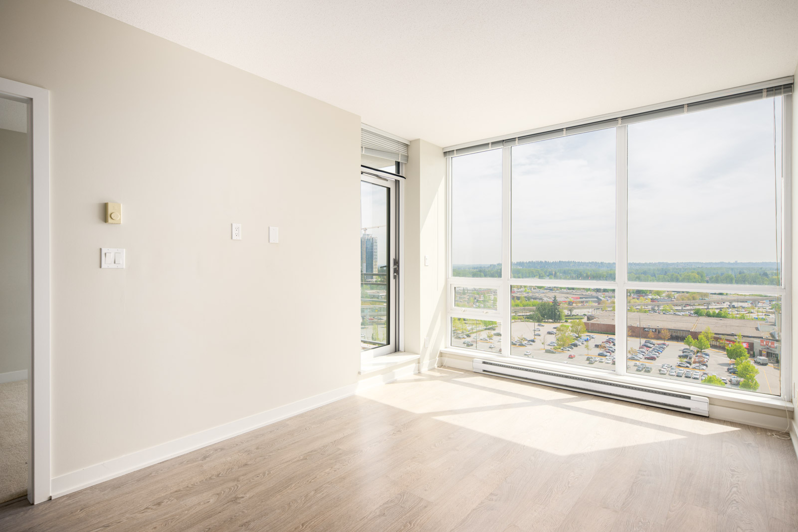 Bright, empty room with light wood flooring, beige walls, and large floor-to-ceiling windows overlooking a parking lot and distant trees.