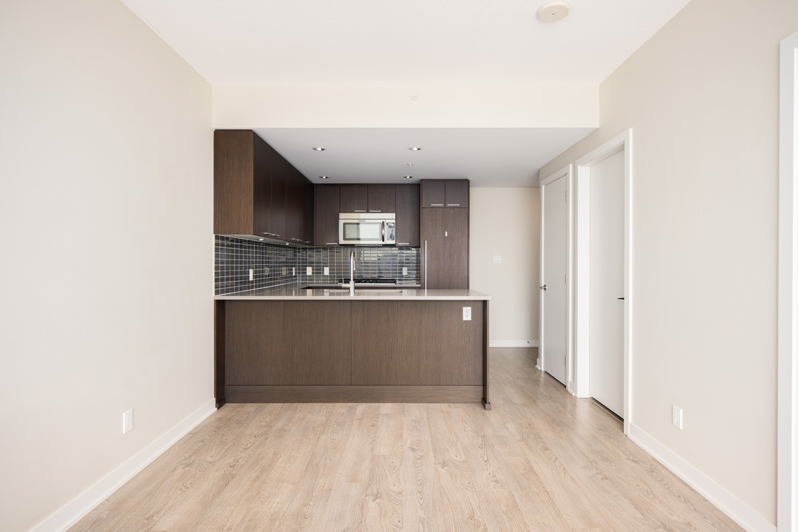Modern kitchen with dark wood cabinets, stainless steel appliances, light wood flooring, and neutral-colored walls in an empty apartment.