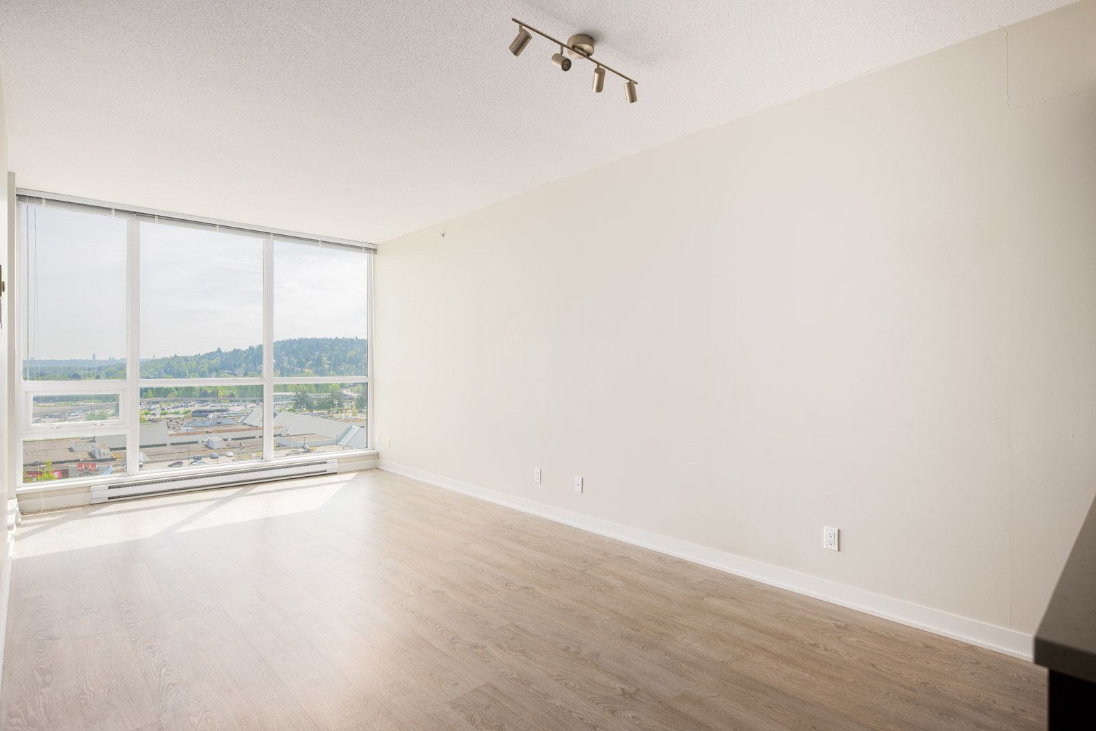 Unfurnished room with light wood flooring, large floor-to-ceiling windows, white walls, and a ceiling light fixture, overlooking a cityscape and hills.