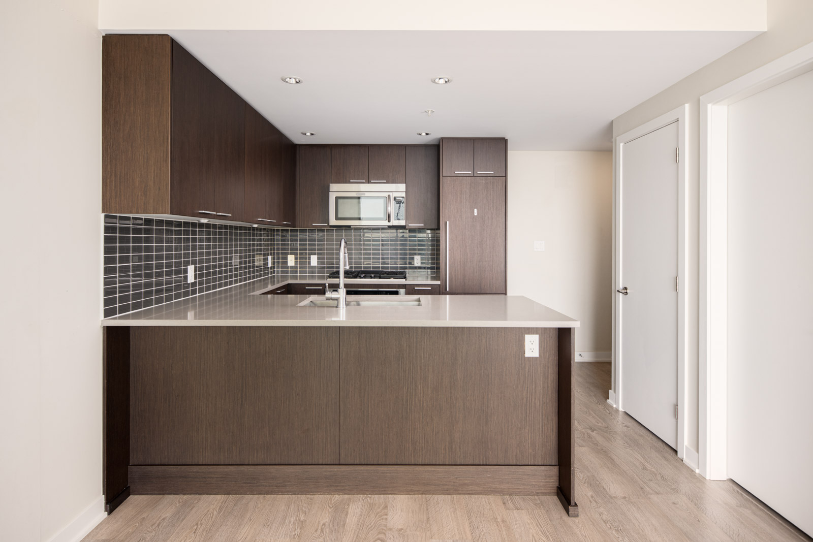 Modern kitchen with dark wood cabinets, gray tile backsplash, stainless steel appliances, and a large island with a sink; light wood flooring throughout.