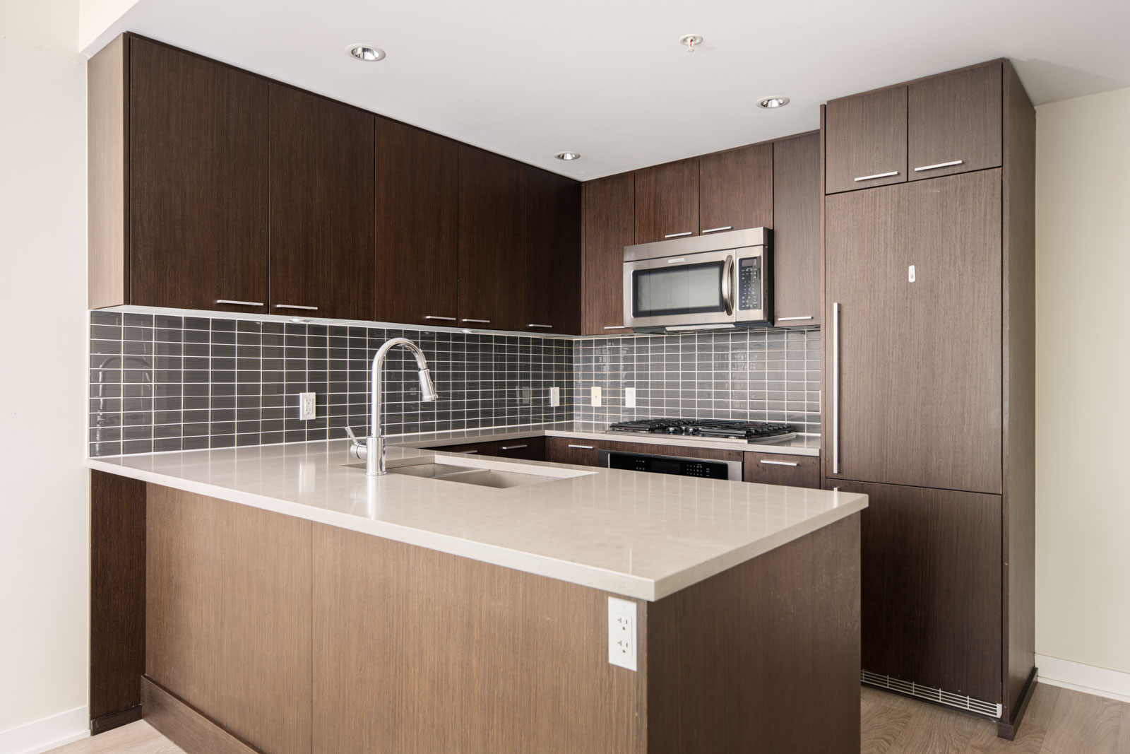 Modern kitchen with dark wood cabinets, gray tile backsplash, built-in stainless steel appliances, and a white countertop with an under-mount sink and gooseneck faucet.