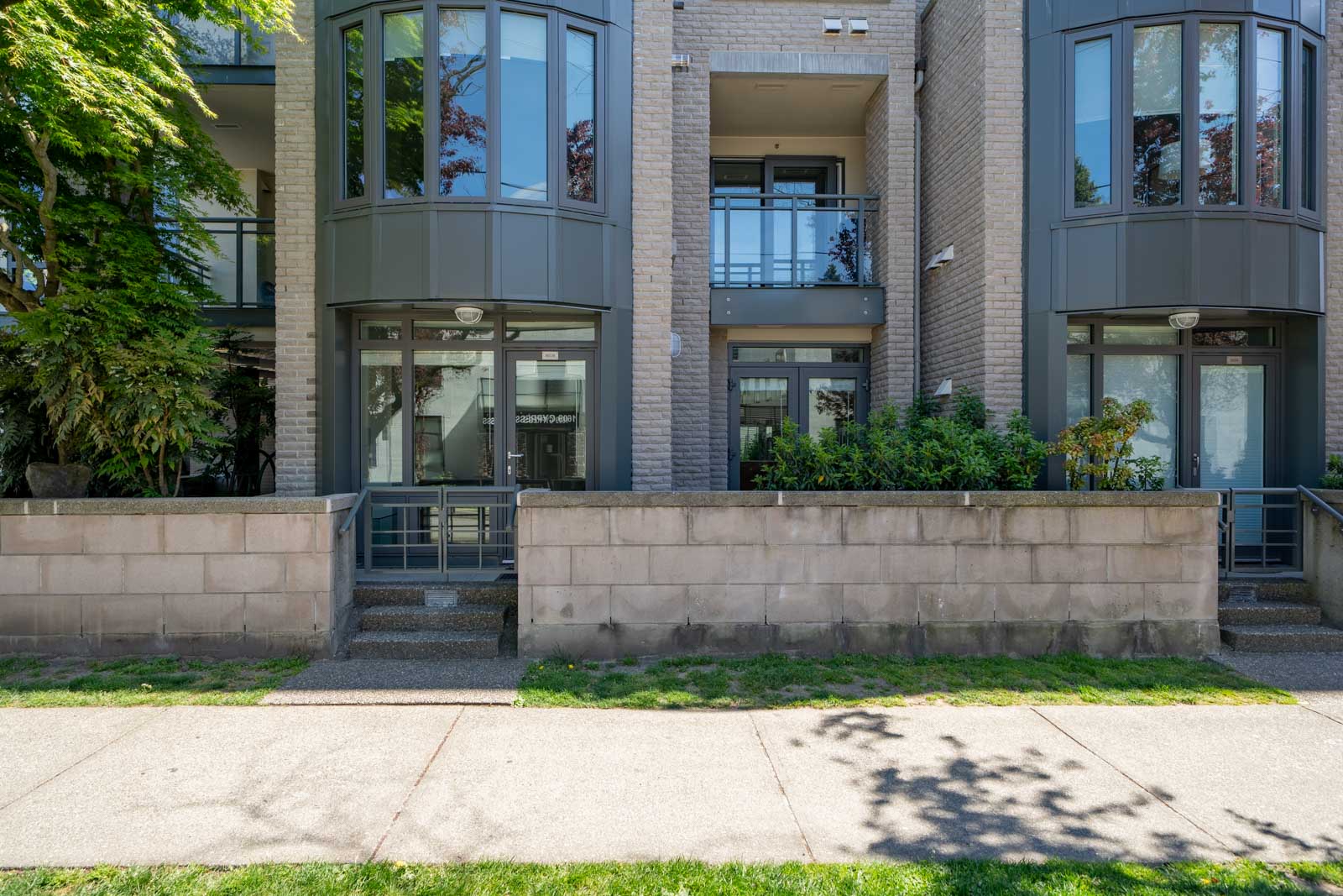 Modern apartment building with large windows, balconies, and a low concrete wall next to a sidewalk and grassy area.