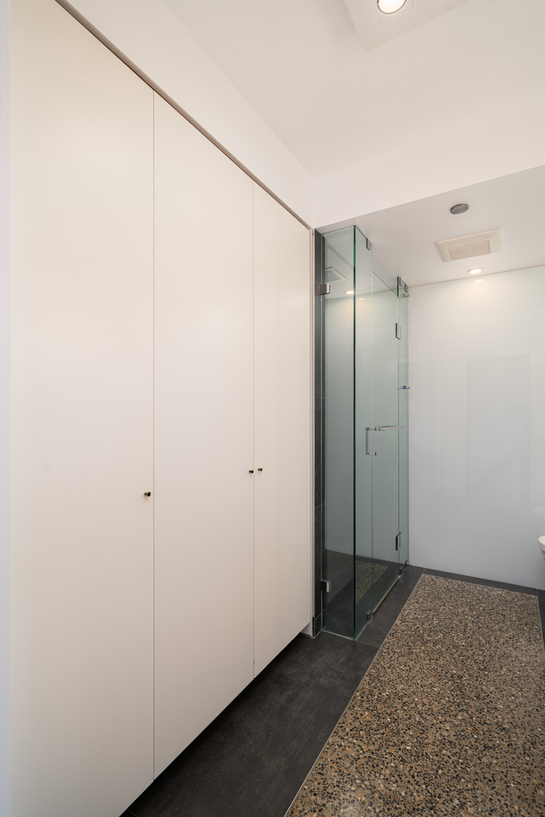 Minimalist bathroom with white cabinets on the left, a glass-enclosed shower, and a speckled stone floor.