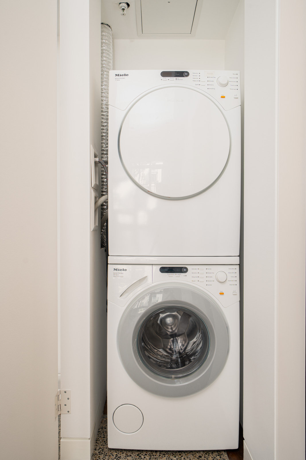 A stacked white washer and dryer unit is installed in a small, narrow laundry closet with light-colored walls.