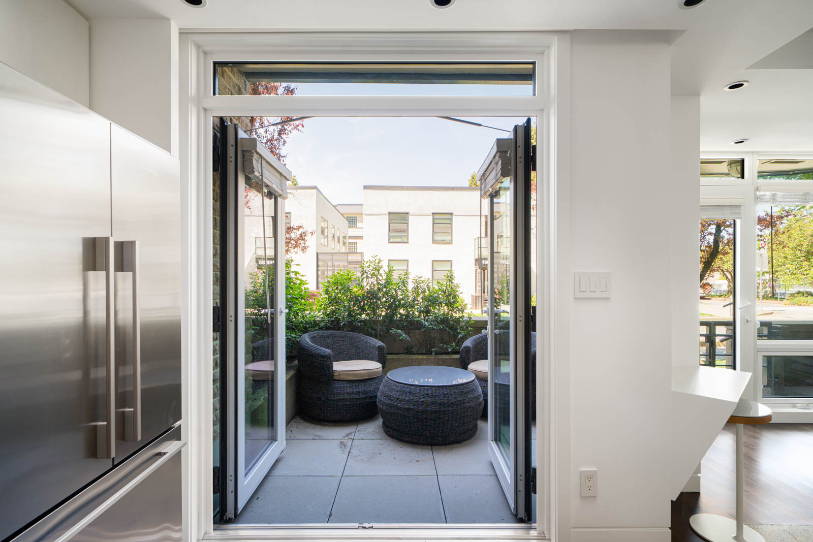 Open glass doors lead from a modern kitchen to a small outdoor patio with round wicker chairs, a table, and greenery, with buildings visible in the background.