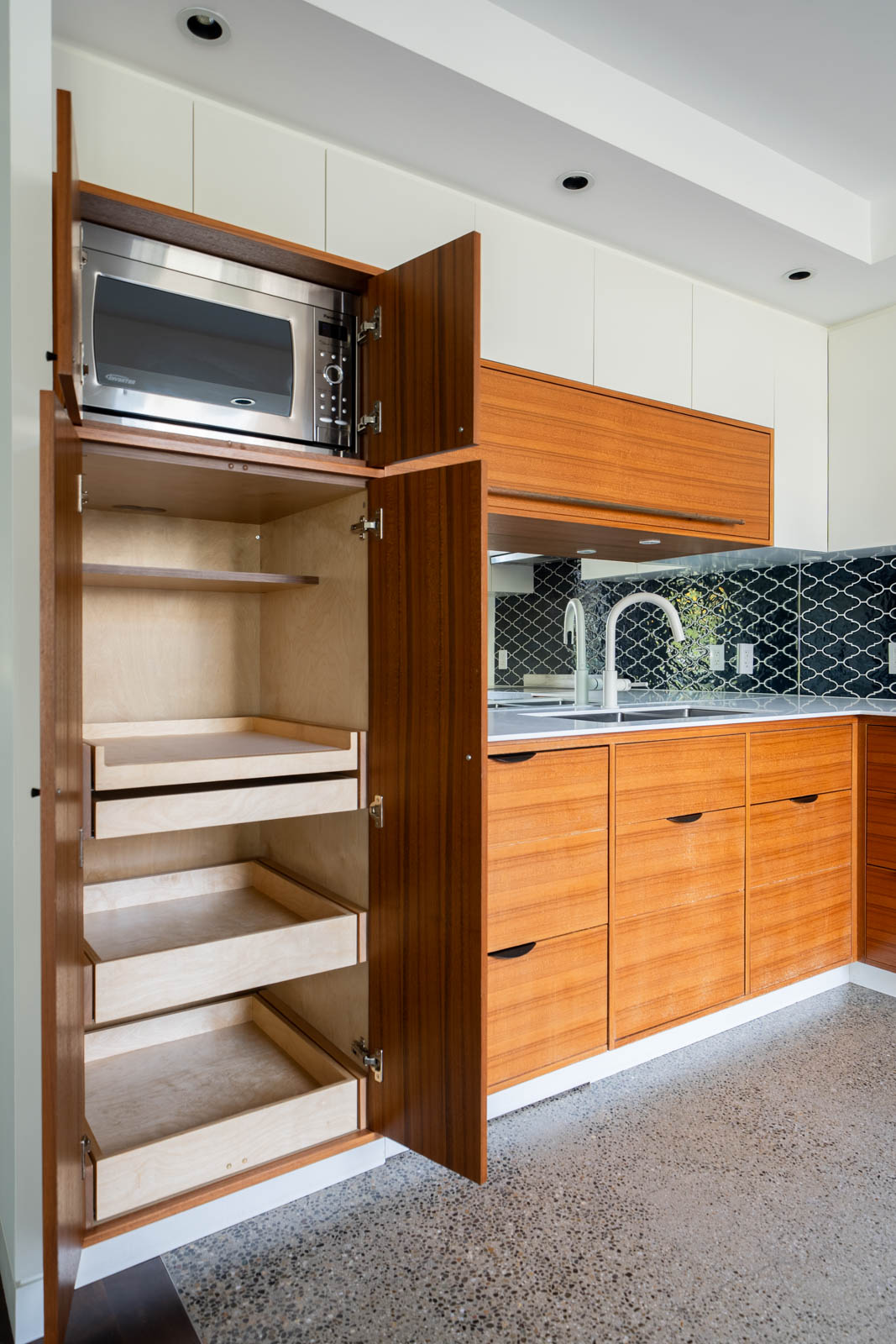 Modern kitchen with open wooden cabinet showing pull-out shelves and a microwave, next to a sink and tiled backsplash.
