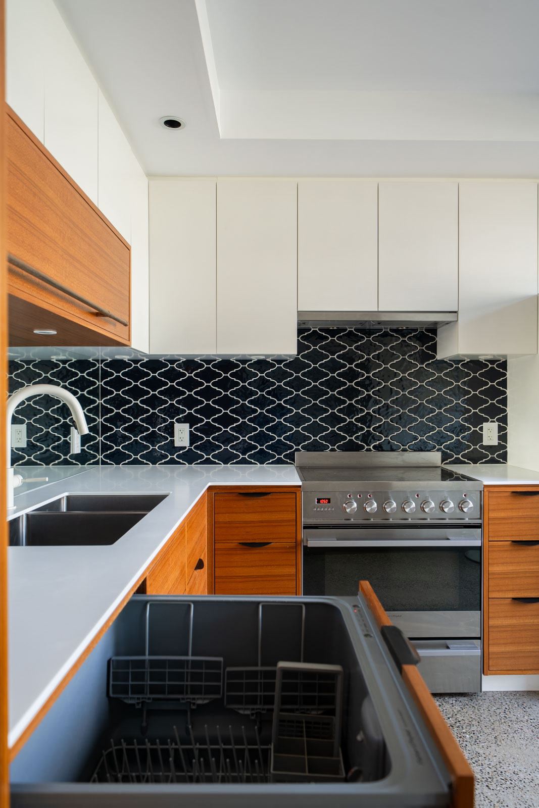 Modern kitchen with wooden cabinets, white upper cupboards, a black patterned tile backsplash, stainless steel oven, and an open dishwasher in the foreground.