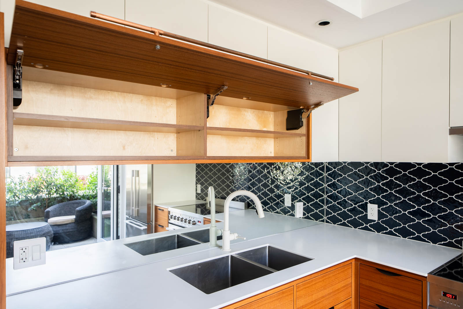 Modern kitchen with an open upper cabinet, white countertops, double sink, black patterned backsplash, and a view of outdoor seating through glass doors.
