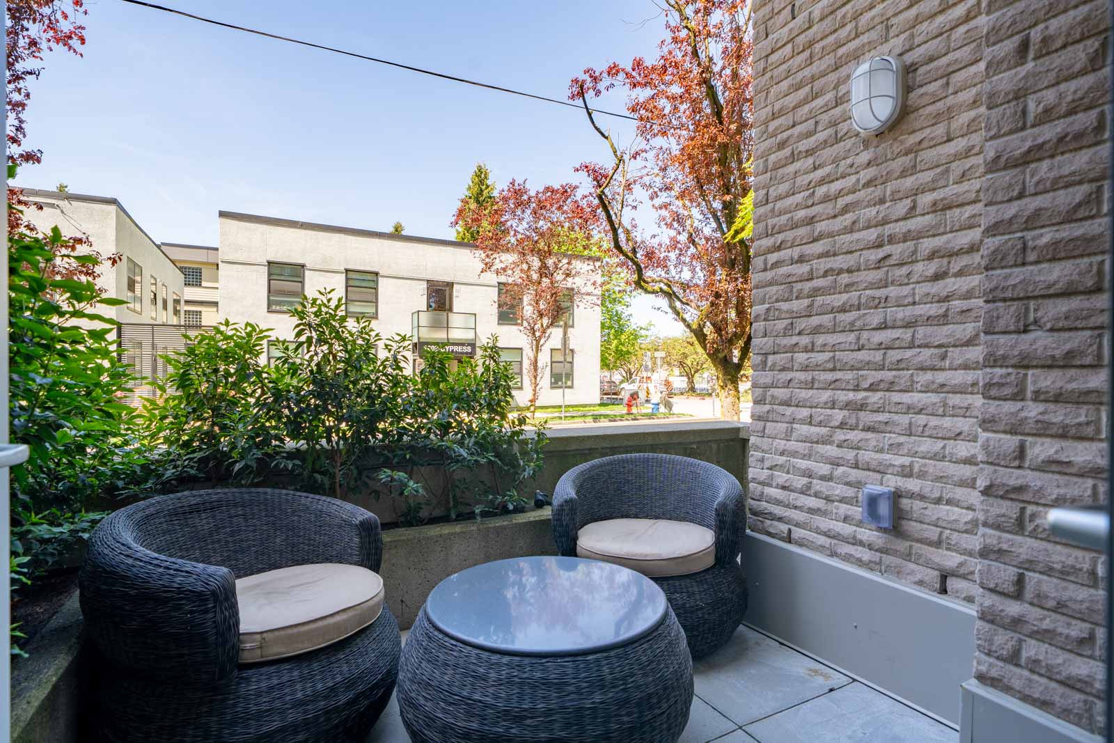 A small outdoor patio with two wicker chairs and a round glass table, surrounded by a low wall and greenery, overlooking a street with modern buildings and trees.