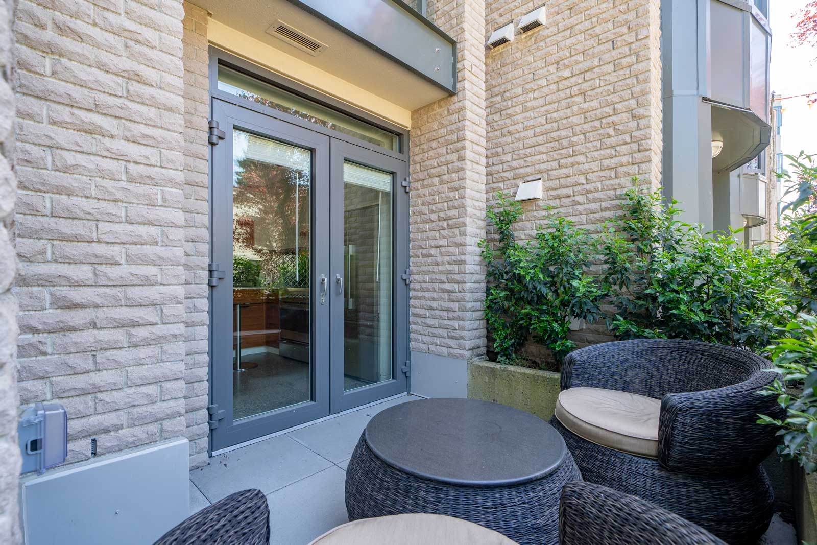 Small outdoor patio with a round table and two wicker chairs with cushions, adjacent to double glass doors and surrounded by beige brick walls and green plants.